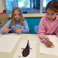 Zwei Mädchen sitzen vor zwei leeren Blättern, halten ihre Bleistifte in der Hand und betrachten ein Kunstwerk.